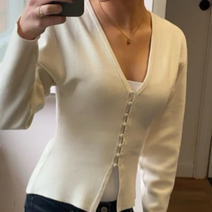 Vit kofta med knappar och v-ringning - Säljer en stilren vit kofta med v-ringning och dekorativa knappar framtill. Koftan har lång ärm och en figurnära passform som ger en clean och fräsch look. Perfekt att matcha med jeans eller kjol för en enkel men snygg outfit.
