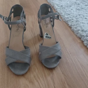Grå sandaler med klack från Duffy - Snygga sandaler  från Duffy med breda korsade remmar framtill och justerbart spänne vid ankeln. Skorna har öppen tå och stabil blockklack som ger extra höjd utan att tumma på komforten. Perfekta för dig som vill ha en stilren och trendig look.