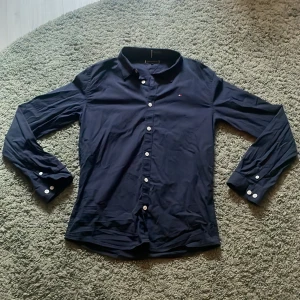 Mörkblå skjorta från Tommy Hilfiger - Priset är ej hugget i sten. Aldrig använd. Snygg mörkblå skjorta från Tommy Hilfiger med klassisk krage och vita knappar framtill. Skjortan har lång ärm och diskret broderad logga på bröstet. Tillverkad i mjuk bomull som känns skön mot huden. Perfekt för dig som gillar stilrena och tidlösa plagg.