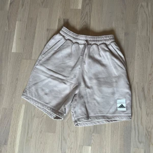 Beige shorts från Adidas - Snygga beige shorts från Adidas med elastisk midja och snörning för perfekt passform. Adidas-logga på ena benet ger en clean och sportig vibe. 