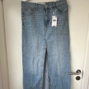 Ljusblå lång jeanskjol med slits - Lång jeanskjol i ljusblå denim med klassiska fickor fram och bak, bälteshällor och en snygg slits framtill. Perfekt för dig som gillar en avslappnad men trendig look. Rå kant nertill för en extra cool vibe.