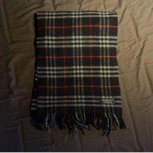 Rutig ullhalsduk från Burberrys - Snygg rutig halsduk från Burberrys i 100% lammull. Färgerna är svart, vit och röd, med klassiskt rutmönster och fransar i ändarna. Perfekt accessoar för att lyfta din outfit med en tidlös brittisk vibe.