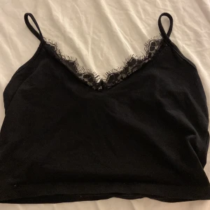 Svart linnetopp med spets från Even&Odd - Svart linnetopp från Even&Odd med smala axelband och snygg v-ringning dekorerad med spetsdetaljer. Perfekt att styla lager på lager eller som den är för en clean look. Mjuk och skön känsla tack vare bomullsmaterialet.