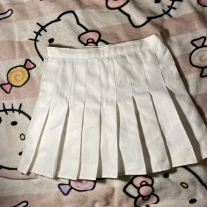 Vit plisserad kortkjol med shorts - Superfin vit plisserad kortkjol med inbyggda shorts under för extra komfort. Kjolen har klassiska veck och stängs med knapp i sidan. Perfekt för en söt och sportig look, funkar lika bra till sneakers som boots.