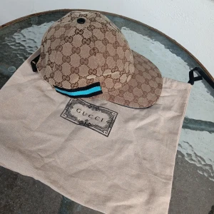 Gucci keps med GG-mönster, beige - Snygg keps från Gucci i beige och brunt med klassiskt GG-monogram över hela kepsen. Har en blå och svart randig detalj på sidan och justerbar rem bak. Perfekt för dig som vill sticka ut med lyxig streetstyle.