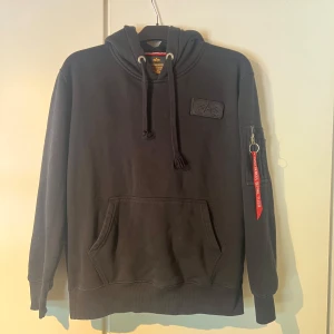 Svart hoodie Alpha Industries M - Svart hoodie från Alpha Industries med röd detalj och text på huvan. Klassisk känguruficka, snörning i halsen och ikonisk 'Remove Before Flight'-tag på ärmen. Badge på bröstet och ärmficka med dragkedja. Perfekt streetwear-vibe. Andvänd fåtal gånger. Nypris ca 1000kr
