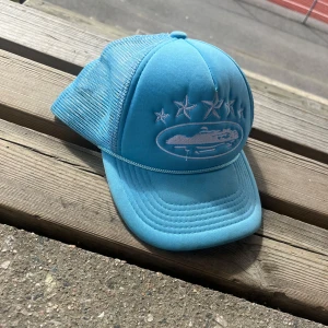 Ljusblå Corteiz keps med brodyr - Säljer en ljusblå corteiz med mesh på sidorna och justerbar snapback. Framsidan har broderade stjärnor och ett ovalt motiv. Perfekt för dig som vill sticka ut med en cool och sportig look.