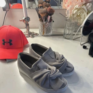 Grå slip-on sneakers med knutdetalj - Snygga grå slip-on sneakers med en cool knutdetalj framtill. Skorna har platt vit sula och är tillverkade i mjuk textil som ger en skön känsla. Perfekta för dig som vill ha en stilren men ändå unik look till vardags.