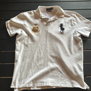 Vit pikétröja Polo Ralph Lauren - Vit pikétröja från Polo Ralph Lauren med klassisk krage, broderad logga i svart och ett snyggt emblem på bröstet. Slim fit-modell i mjuk bomull, perfekt för en clean och stilren look. Kortärmad och riktigt fräsch till sommaren.