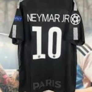 PSG Neymar Jr svart fotbollströja - Svart PSG fotbollströja med Neymar Jr och nummer 10 på ryggen. Tröjan har korta ärmar, vita detaljer och tryck, samt Champions League-märke på ärmen. Perfekt för dig som älskar fotboll och vill visa ditt stöd för Neymar och Paris Saint-Germain.