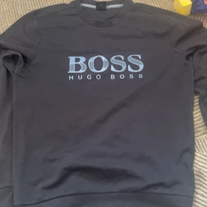 Svart sweatshirt från Hugo Boss - Svart sweatshirt från Hugo Boss med stort BOSS-tryck i ljusblått på bröstet. Klassisk rund halsringning och långa ärmar. Tröjan är i mjuk bomull och har en clean, stilren look som passar perfekt till jeans eller joggers.