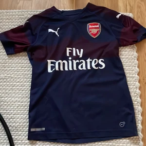 Arsenal fotbollströja - Snygg Arsenal fotbollströja från Puma i marinblått med vinröda detaljer på axlarna. Tröjan har korta ärmar, Arsenal-emblem på bröstet och Fly Emirates-tryck framtill. perfekt för fritid eller träning.