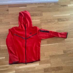 Röd Nike hoodiejacka med dragkedja - Nike hoodiejacka i rött med svarta detaljer och hel dragkedja. Jackan har huva, svarta linjer på bröstet och en ficka med dragkedja på ena ärmen. Perfekt för dig som gillar sportig och avslappnad stil.