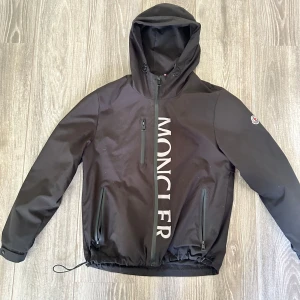 Svart vindjacka från Moncler med huva - Svart vindjacka från Moncler i storlek M med stor logga framtill, huva med dragsko och tre dragkedjefickor. Jackan har Moncler-märke på ärmen och justerbar nederkant. Liten repa syns på sista bilden.