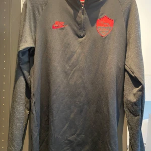 Svart långärmad AS Roma Nike Dri-FIT - AS Roma långärmad träningströja från Nike i svart Dri-FIT-material. Tröjan har röd AS Roma-logga och Nike Swoosh på bröstet, halv dragkedja framtill och diskret röd detalj i nacken. Perfekt för fotbollsträning eller supporterstil.