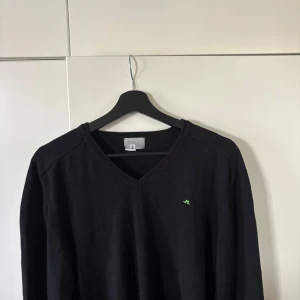 Svart v-ringad tröja från J.Lindeberg - Svart v-ringad tröja från J.Lindeberg med diskret grön logga broderad på bröstet. Tröjan är i mjuk ull och har långa ärmar samt ribbade muddar vid hals, ärmslut och nederkant. Perfekt för dig som gillar stilren och enkel design.