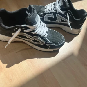 Svarta B30 sneakers med vita detaljer - Snygga svarta B30 sneakers med vita och grå detaljer samt tydlig CD-logga på sidan. Skorna har vit sula och vita snören, och är tillverkade i textil och syntetmaterial. Perfekta för dig som vill sticka ut med exklusiva och trendiga sneakers.