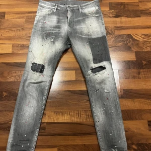 Grå Dsquared2 jeans med färgstänk - Snygga grå jeans från Dsquared2 med slitna detaljer, hål på knäna och coola färgstänk i vitt och rött. Modellen har en smal passform och klassisk femficksdesign. Stort Dsquared2-märke bak i midjan och logotyp vid gylfen. Perfekta för dig som gillar streetstyle.