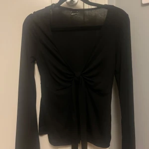 Svart topp från Gina Tricot - Svart långärmad topp med knytning vid bröstet  och djup v-ringning. Den är öppen vid magen också. Toppens material är mjukt, stretchig och luftigt. Aldrig använd bara provat på!