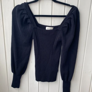 Svart ribbad tröja Second Female - Supersnygg svart ribbad tröja från Second Female med markerade axlar och långa ärmar. Tröjan har en fyrkantig ringning och breda muddar vid ärmsluten. Perfekt till jeans eller kjol för en trendig look.