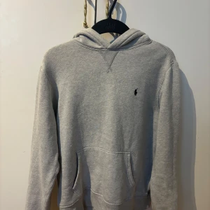 Grå hoodie från Ralph Lauren - Klassisk grå hoodie från Ralph Lauren med broderad logga på bröstet. Tröjan har huva, känguruficka och långa ärmar. Perfekt för en chill och avslappnad stil. Passar folk runt 160. 