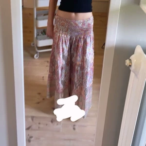 Mönstrad långkjol från Marta Du Chateau - Superfin långkjol med färgglatt blommigt mönster i rosa, beige och orange toner. Kjolen har smock i midjan för en bekväm passform och är gjord i ett lätt och luftigt material, perfekt för varma dagar. Snygg att styla med en enkel topp eller stickad tröja.