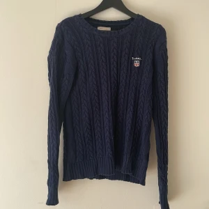 Mörkblå  tröja från  Bondelid😍 - Kabelstickad mörkblå tröja från Bondelid med broderad logga och sköld på bröstet. Tröjan har rund halsringning, långa ärmar och ribbade muddar vid ärmslut och nederkant. Perfekt för dig som gillar klassisk och stilren look.