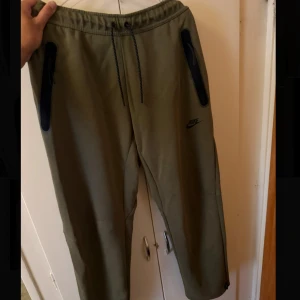 Olivgröna mjukisbyxor från Nike - Snygga olivgröna mjukisbyxor från Nike med svarta dragkedjor på fickorna och justerbar midja med snörning. Perfekta för chill eller träning, med en modern och avslappnad passform. Nike-logga på låret för extra stilpoäng.