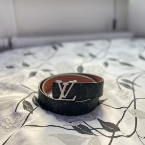 Louis Vuitton Bälte - Snyggt svart bälte från Louis Vuitton med klassiskt monogrammönster och stor LV-logga i silver på spännet. Bältet är gjort i skinn och har en brun insida. Perfekt accessoar för att lyfta din outfit med en lyxig touch. Storlek 115cm