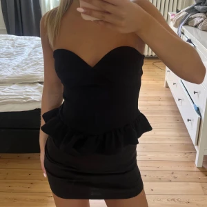 Svart axelbandslös volang topp Object - Supersnygg svart axelbandslös topp från märket Object, köpt på Vero Moda. Volangdetalj i midjan och figurnära passform. Klänningen har en hjärtformad urringning och är perfekt för dig som vill sticka ut med en stilren look.