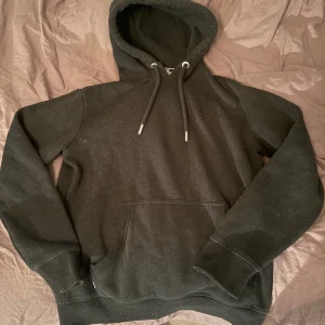 Svart hoodie från SuperDay - Mörkgrå hoodie från SuperDay med broderad logga på bröstet. Klassisk modell med huva, dragsko och stor magficka. Mjuk insida och ribbade muddar vid ärmslut och nederkant. Perfekt för chill dagar eller när du vill ha en enkel streetstyle.