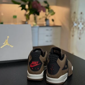 Nike Air Jordan 4 Travis Scott Cactus Jack - Nike Air Jordan 4 Travis Scott Cactus Jack i brun mocka med svarta och beige detaljer. Snygg röd Jumpman-logga på plösen och Nike Air-brodyr bak. Svart snörning, vit och svart sula med röda detaljer undertill. Unik tag med Cactus Jack-tryck. Perfekt för dig som vill sticka ut.