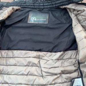 Moose Knuckles väst - Snygg svart dunväst från Moose Knuckles med quiltad design och dragkedja framtill. Västen har hög krage, två fickor med dragkedja och diskret logga på bröstet. Perfekt för lager-på-lager och håller dig varm under kyliga dagar. Storlek L men passar också M