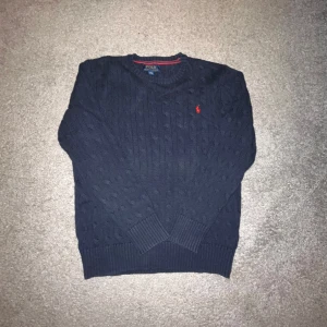 Stickad Polo Ralph Lauren tröja - Säljer en marinblå kabelstickad Polo Ralph Lauren tröja som är sparsamt andvän. Storlek 14/16 år men skulle säga att den är väldigt liten i storleken. Hör av dig vid minsta fundering.