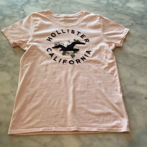 Ljusrosa Hollister t-shirt med brodyr - Fin ljusrosa t-shirt från Hollister med broderad logga och blommor framtill. Klassisk rund halsringning och korta ärmar. Mjuk bomullskvalitet. Storlek xs men känns mer som en s 💕 