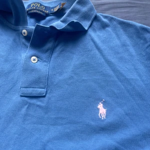 Blå pikétröja från Polo Ralph Lauren - Snygg blå pikétröja från Polo Ralph Lauren med klassisk krage, två knappar och den ikoniska broderade loggan i vitt på bröstet. Tröjan är i bomull och har korta ärmar, perfekt för en clean och sportig look.