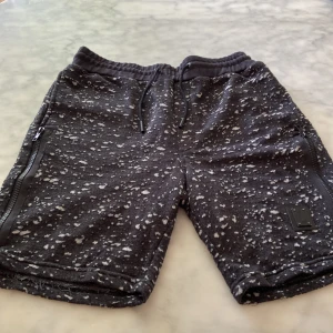 Svarta shorts från Religion med dragkedjor - Coola svarta shorts från Religion Clothing med vitt spräckligt mönster. De har elastisk midja med snörning, två dragkedjor på sidorna och en Religion Clothing -logga på benet. Perfekta för chill dagar eller streetwear. Mjukt material som känns skönt mot huden.