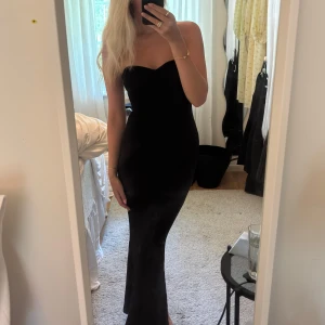 Svart axelbandslös långklänning - Supersnygg svart axelbandslös långklänning med figurnära passform och lätt utsvängd nederdel. Klänningen har en enkel och elegant design med hjärtformad urringning och är perfekt för dig som vill ha en stilren look.