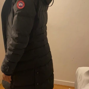 Svart lång dunjacka från Canada Goose - Säljer en svart lång dunjacka från Canada Goose med huva och snygg pälsdetalj. Jackan har klassisk patch på ärmen, dragkedja och knappar framtill. Perfekt för kalla dagar och riktigt varm. Jag har haft den i några år och har bara använt den i en vinter. Jackan är äkta och kvitto finns vid intresse.  Priset kan diskuteras 