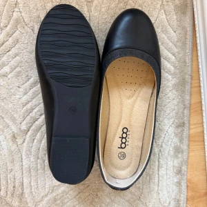 Svarta ballerinaskor från Bobo Shoes - Klassiska svarta ballerinaskor från Bobo Shoes med rund tå och platt sula. Skorna har en enkel och stilren design i syntetmaterial med mjuk innersula för extra komfort. Perfekta för dig som gillar en clean och tidlös look. Aldrig använda