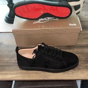 Svarta sneakers från Christian Louboutin - Snygga svarta sneakers från Christian Louboutin i mocka med klassisk röd sula. Skorna har rund tå, svarta skosnören och diskret design som ändå sticker ut med den ikoniska röda undersidan. Perfekta för dig som vill ha en lyxig touch till din outfit.