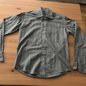 Randig grå skjorta från Robert Rossi - Snygg grå skjorta från Robert Rossi med tunna vita och ljusbruna ränder. Klassisk krage, långärmad och med knappar framtill samt bröstficka. Skjortan har en stilren look och passar perfekt till dig som gillar en clean och enkel stil.