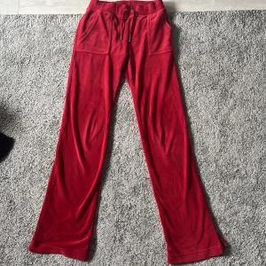 Röda mjukisbyxor från Juicy Couture - Snygga röda mjukisbyxor från Juicy Couture i mjuk velour med bred resår i midjan och snörning. Byxorna har stora fickor fram och bak med silverfärgade knappar med logga. Perfekta för en chill och trendig look.