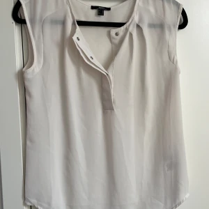 Vit ärmlös blus från Mexx - Säljer en stilren vit blus från Mexx i storlek 36. Blusen är ärmlös med en lätt transparent känsla och har knappar framtill vid halsringningen. Perfekt för en clean och fräsch look, passar till både jeans och kjol.