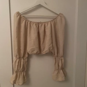 Beige offshoulder blus från Trendyol - Supersöt beige offshoulder blus från Trendyol med vida, volangiga ärmar och smockdetaljer. Toppen är croppad och har en luftig känsla, perfekt för en chill och trendig look. Passar dig som gillar romantiska vibes och vill sticka ut lite extra.