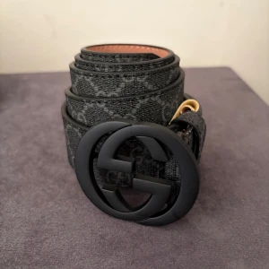 Gucci Bälte med GG märke - Snyggt svart bälte från Gucci med klassiskt GG-mönster och stor rund mattsvart GG-spänne. Utsidan är mönstrad i mörkgrått och svart, insidan är ljusbrun skinn. Perfekt accessoar för att lyfta din outfit med lyxig känsla och ikonisk design.