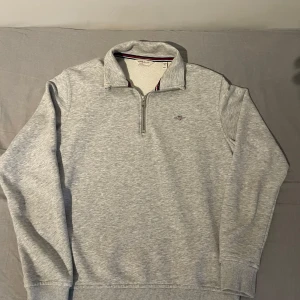 Grå half zip tröja från GANT - Snygg ljusgrå tröja från GANT med halv dragkedja och broderad logga på bröstet. Tröjan har ribbade muddar och krage med randig insida. Perfekt för dig som gillar klassisk och stilren look.