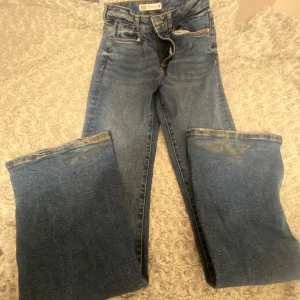 Blå bootcut jeans från Zara - Säljer ett par klassiska blå bootcut jeans från Zara med fem fickor och snygga kontrastsömmar. Jeansen har hög midja och är tillverkade i ett robust bomullstyg. Utsvängda också. Perfekta för dig som gillar en tidlös och avslappnad look.