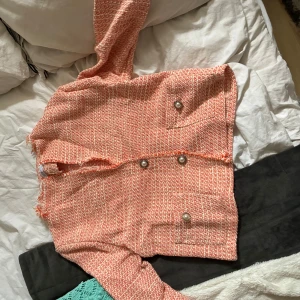 Tweed kavajjacka från Hollies XS - Snygg tweed kavajjacka från Hollies i orange och vitt och rosa. Jackan har dekorativa pärlknappar framtill och två fickor. Den är kortare i modellen och har en klassisk, chic look som passar perfekt till jeans eller kjol.