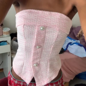 Rosa tubtopp med pärlknappar - Supersöt rosa tubtopp i strukturerat tyg med tre glittriga pärlknappar framtill och snygg spetsig nederkant. Passar perfekt till någon middag, resturang, fest osv :) Toppen har dragkedja i ryggen och är axelbandslös, perfekt för att sticka ut. Passformen är tight och materialet känns lyxigt och lite tjockare. Samt har jag använt denna tip endast en gång! 